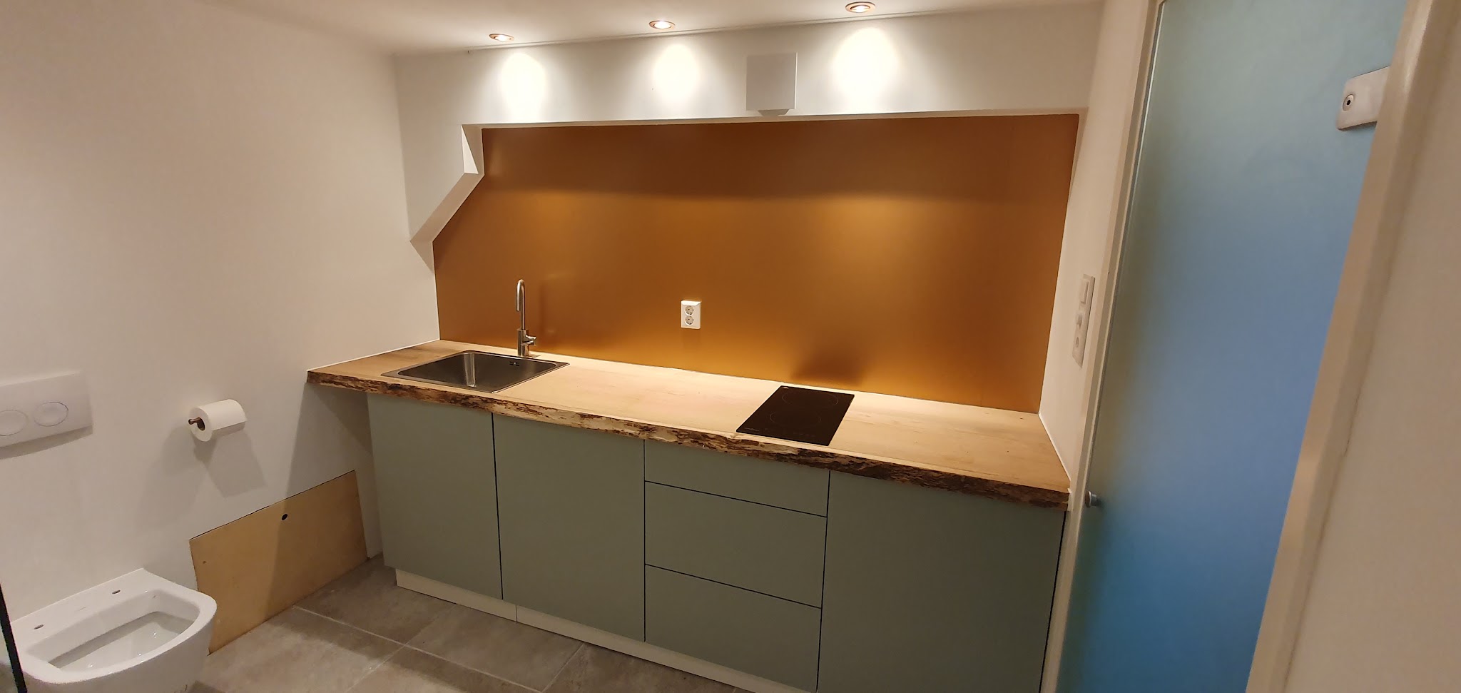 Afgewerkt keukenblok met groene kasten, een houten blad met boomstamrand en een koperkleurige achterwand