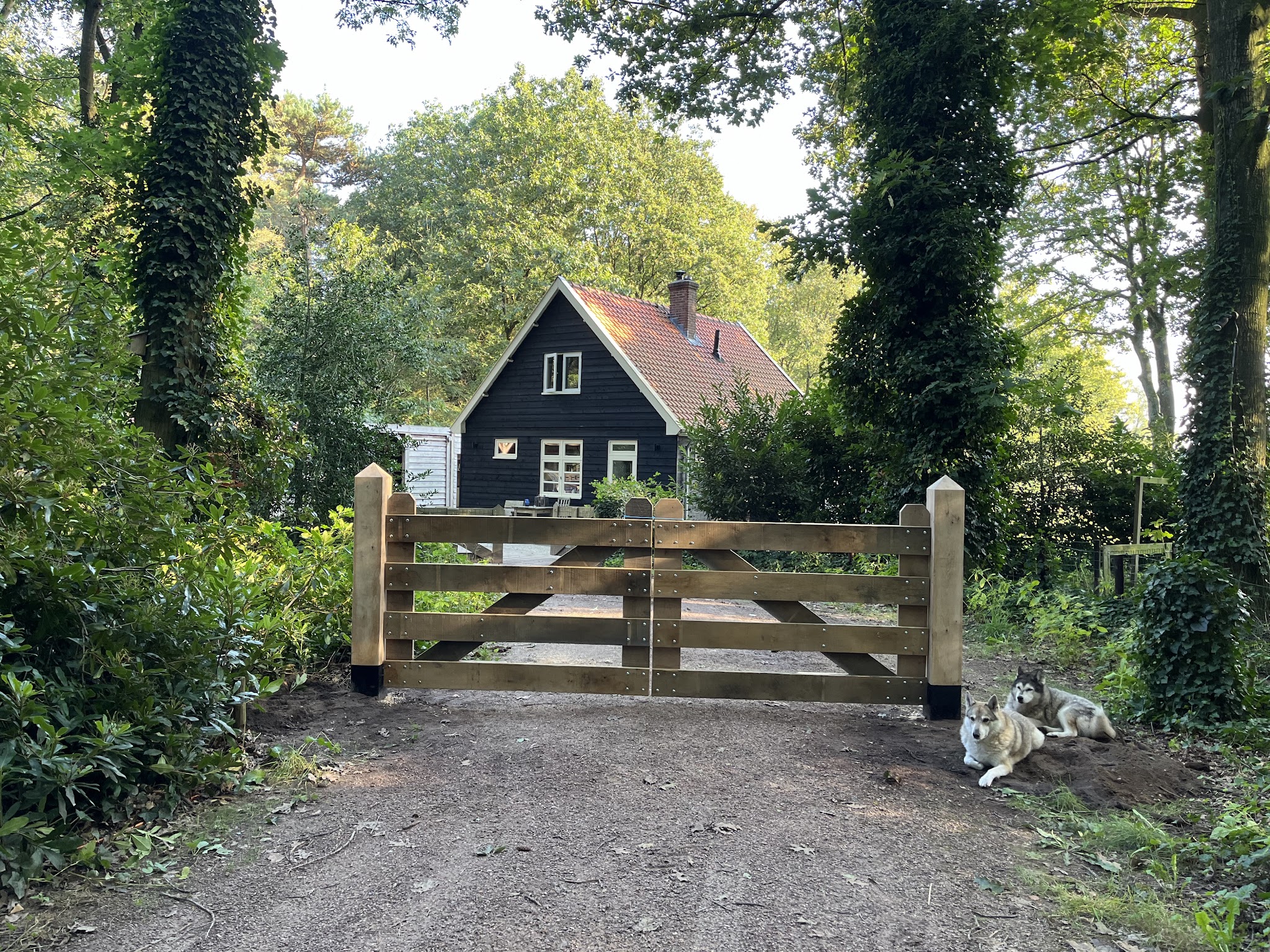 Zelfgemaakte eiken inrijpoort op een bosweg, met een zwart houten huis erachter